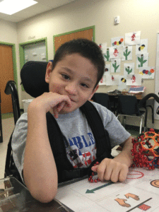 A student in his wheelchair; a universal core communication board is taped to a tray in front of him.