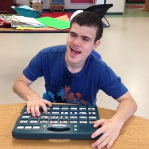 An adult is holding out a universal core board and pointing at a symbol as a student looks over it.